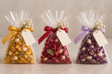 Load image into Gallery viewer, Clear cellophane gift bags filled with dried botanicals and flowers, tied with yellow, red, and purple ribbons and decorated with blank tags, arranged on a light beige surface against a soft neutral background. Party Favours rovistella